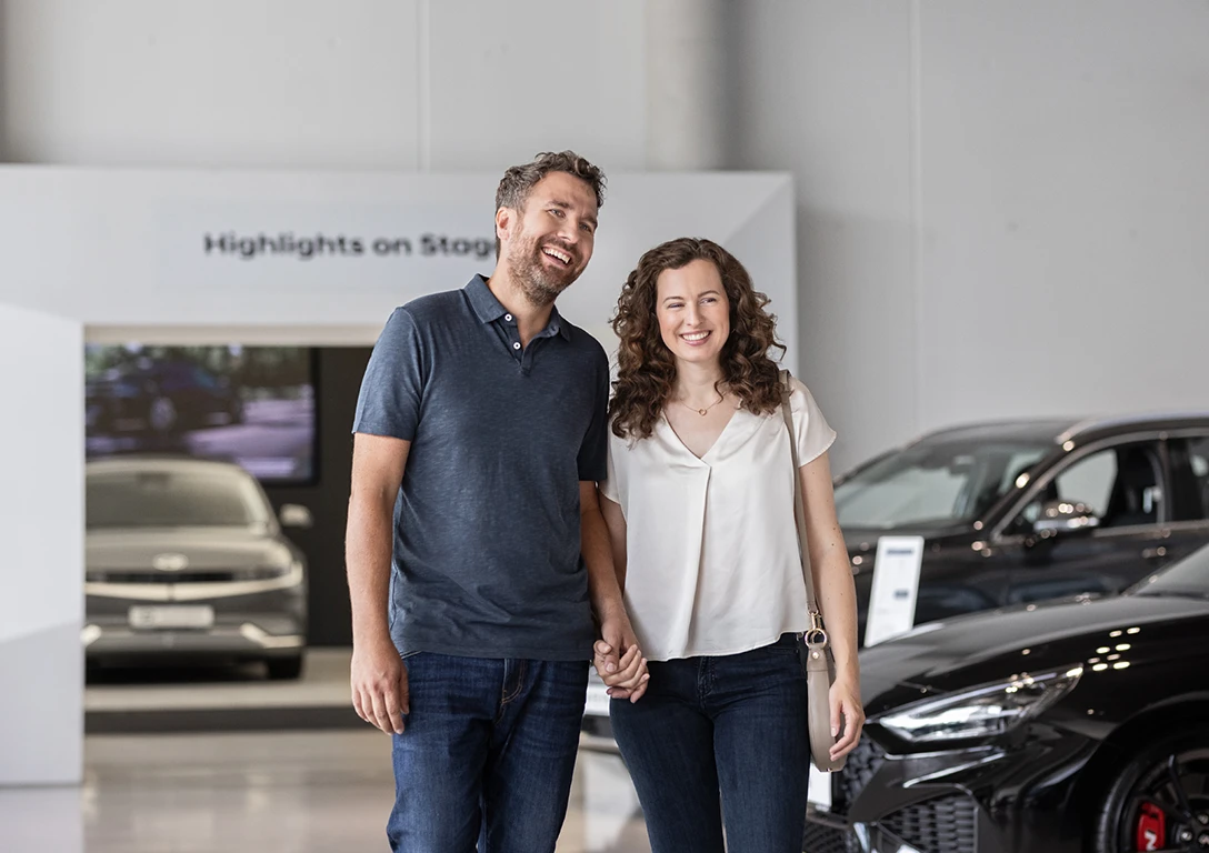 „Lächelndes Paar hält Händchen und geht durch einen hellen Autohaus-Showroom mit mehreren ausgestellten Fahrzeugen.“